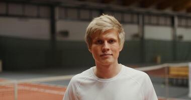 Young handsome blonde male tennis player smiling looking straight at the camera, putting a tennis racket on his shoulder, close-up video
