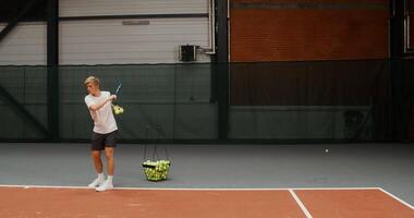 A young man of European appearance, dressed in sportswear, throws tennis balls with a racket over the net one by one, playing alone video