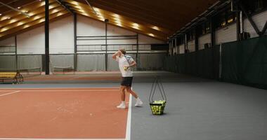 A young handsome man of European appearance with an athletic build, throws tennis balls with a racket over the net one by one, taking them from a basket standing nearby, playing alone video