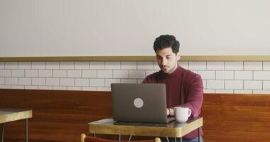 Remote work in a stylish coffee shop over a cup of coffee. A young brunette man, of Armenian appearance, dressed in casual clothes, is typing on a laptop sitting at a table. video