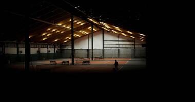 In a large indoor sports hall with several tennis courts, lights are turned on and a man is seen walking towards the bench with a racket and a basket full of tennis balls in his hands. video