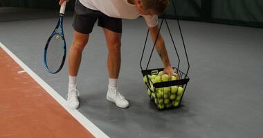 A young man of European appearance takes several tennis balls from a basket standing nearby and alternately hits them with a racket, throwing them on the floor video