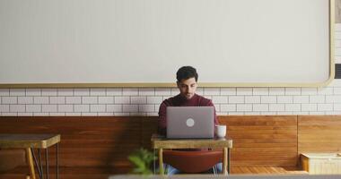 Young dark-haired man with thick stubble on his face, dressed in casual clothes, uses a laptop while sitting at a small table in a cafe, working remotely video
