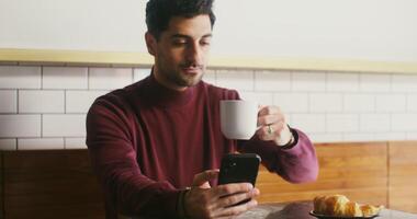 Young black-haired man with a slight unshaven face, dressed in casual clothes, enjoying coffee from a large white mug and using the phone while sitting at a table in a cafe video