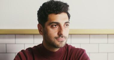 Young black-haired man with a slight unshaven face, dressed in casual clothes, turns his head and smiles broadly, looking directly into the camera video