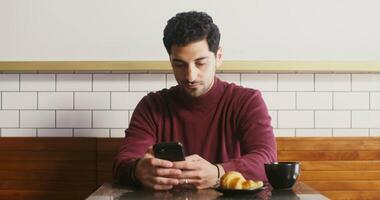 Young Georgian man with stubble on his face, sitting at a small table in a cafe, drinking coffee and typing on the phone, having breakfast alone video