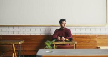 A young dark-haired man with a slight unshaven sits at a small table in a cafe with a mug of coffee and a croissant and uses the phone, from time to time dreamily looking around in anticipation video
