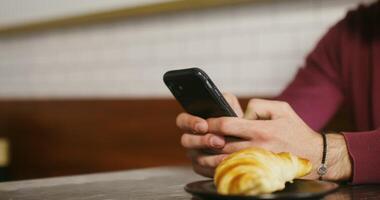 A young man, dressed in casual clothes, uses the phone and drinks coffee while sitting at a small table in a cafe. Close-up footage moving from a man's phone in his hands to a mug video