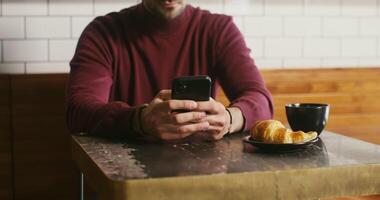 A young man, dressed in casual clothes, uses the phone sitting at a small table in a cafe, ordered a mug of coffee with a croissant. Male hands with a phone close-up, faces are not visible video