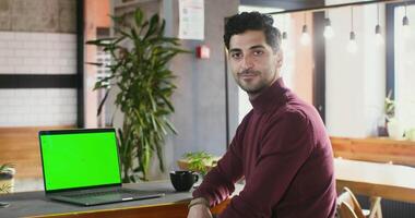 Young brunette man with stubble on his face smiling looking straight into the camera while sitting at a table in a cafe in front of an open laptop with a green screen. Copy space, chroma key video