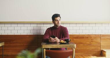 A young brunette man with stubble on his cheeks sits at a small table in a coffee shop with a mug of coffee and a croissant and uses the phone, occasionally looking around dreamily video
