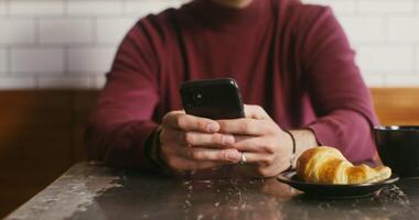 A young man, dressed in casual clothes, uses the phone and drinks coffee while sitting at a small table in a cafe. Male hands close-up, face is not visible video