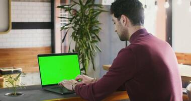 A young brunette man with stubble on his face is concentrating on typing on a laptop with a green screen, sitting at a table in a coffee shop half-turned to the camera. Chroma key, copy space video