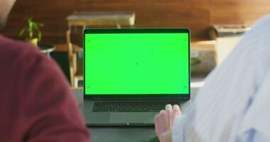 Two people sit at a table in a cafe in front of an open laptop with a green screen. Filming from the back. Chroma key, copy space video
