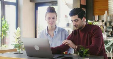 A handsome dark-haired man with stubble on his face is talking to a young beautiful girl of European appearance, while sitting with her at a table in a cafe and showing her a laptop screen video