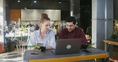 Young beautiful couple smiling and talking while using a laptop together and drinking coffee while sitting at a table in a modern cozy cafe video