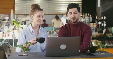 A young Armenian man is typing on a laptop, showing the screen to the girl who is sitting next to him at a table and drinking coffee as they spend time in a stylish cafe talking and laughing video
