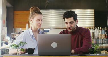 Two young people - a man and a woman laugh and drink coffee while talking to each other and using a laptop, sitting at a table in a cafe. Remote work on a joint project video