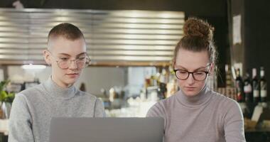 Two modern young women of European appearance communicate with each other while sitting in front of an open laptop in a cozy stylish coffee shop. Online learning, joint project, teamwork video