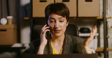 Young beautiful woman of European appearance talking on a mobile with a serious look, sitting in front of an open laptop at a table in a modern stylish office video