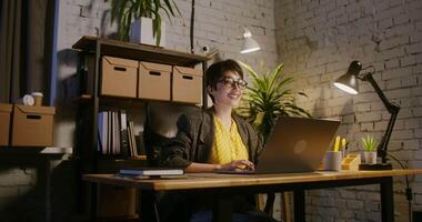 Beautiful young woman of European appearance in glasses, with short haircut, smiles while looking at the camera while sitting at a table in front of an open laptop in a modern stylish office video