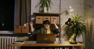 Beautiful young woman of European appearance in glasses emotionally rejoices, raising her hands up and dancing while sitting at the table in front of an open laptop in a modern office video