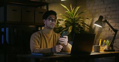 A young brunette man, wearing glasses, dressed in casual clothes, emotionally rejoices after the received SMS and continues typing on the mobile, sitting in front of an open laptop in the office video