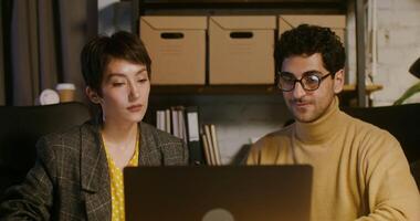 Two young stylish man and woman colleagues work together sitting at a table in front of an open laptop in a modern stylish office. shot from the front video