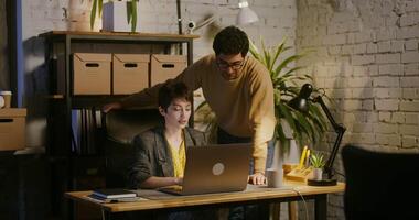 Senior colleague - a young handsome brunette man is teaching a young woman working on a laptop, standing near her and pointing at the screen. Internship in a modern stylish office video