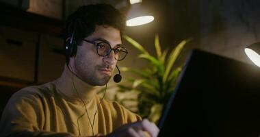 Young dark-haired man with glasses, with stubble on his cheeks, talking on a microphone in headphones while sitting in front of an open laptop under the light of lamps video