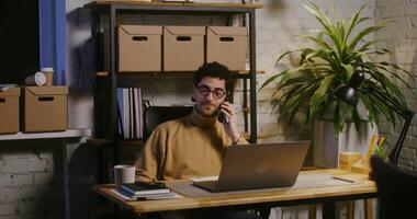 Young handsome curly haired brunette man, wearing glasses talking on a mobile phone while sitting in front of an open laptop in a modern stylish office video