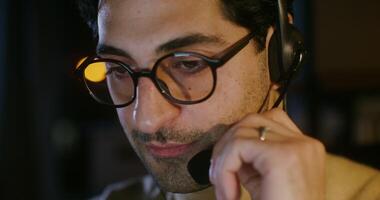 A young dark-haired man with glasses, with stubble on his cheeks, talking on a microphone in headphones, adjusting the microphone with his hand, close-up. Filming in the dark video