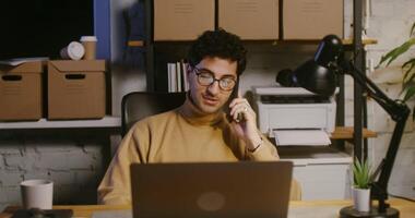 Young handsome curly haired brunette man, wearing glasses talking on a mobile phone and using a laptop, working in an office at night under artificial lighting video