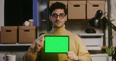 A young handsome curly haired brunette man, wearing glasses, looks directly into the camera, shows a tablet with a green screen and speaks. Copy space, screen sharing video