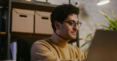 Young handsome curly dark-haired Jewish man answers a mobile phone call and starts talking while sitting in front of an open laptop, in a modern stylish office video
