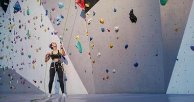 A young woman pulls on a safety rope for a man descending from a climbing wall while standing at the foot of the wall. limbing workout on an artificial surface in the indoor gym video