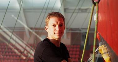 A young man of European appearance is smiling looking at the camera while standing on a climbing wall and holding a safety hook. Climbing classes in the indoor gym video