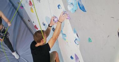 A young man of European appearance climbs up artificial surface of a climbing wall, a young woman belays him while standing at the foot of the wall and holding a safety cable, shoot from above video