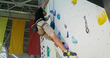A young man is engaged in rock climbing on an artificial surface in an indoor climbing wall, going down the ledges. Climbing equipment for climbing to the top video