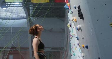 Young beautiful woman athlete, dressed in a sports top, with a safety belt at the waist, looks up while standing in front of a climbing wall, about to climb video