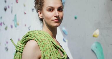 Young beautiful woman athlete with prepared safety ropes on her shoulder looking straight into the camera with a serious look, standing near the climbing wall and getting ready to climb video