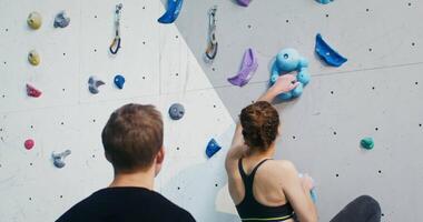 A man of European appearance stands at the foot of the climbing wall, and watches as a young woman athlete climbs up the artificial stones and fixes the rescue rope. video