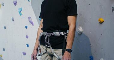 Young man of European appearance wearing a safety belt, smiling looking straight into the camera, standing in front of a climbing wall in an indoor hall, dynamic shooting video