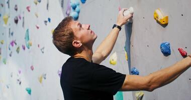 A man of European appearance, equipped with safety cables hanging from a strap, stands at the foot of the climbing wall and looks up, thinking over a route for climbing video