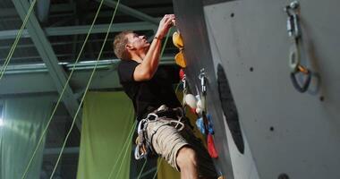 A young man of European appearance in rubberized sports shoes, wearing a safety belt, with a serious look, climbs up the climbing wall, treating his hands with talcum powder on the weight video
