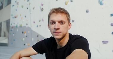 A young man of European appearance sits on the floor near an climbing wall and smiles while looking into the camera. Portrait videography in the gym under artificial lighting video