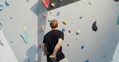 A man of European appearance treats his hands with talcum powder, dropping them into a bag hanging on a belt behind his back, and begins to climb the climbing wall without belay. Filming from the back video