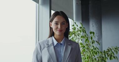 Young beautiful woman of European appearance, dressed in a business suit, smiling looking at the camera, standing near the window in a modern office video