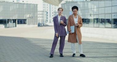 Two business women of different nationalities with glasses with coffee in their hands are looking at the camera, standing near the modern business center, on a sunny autumn day video