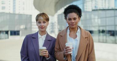Two business women of different nationalities with glasses with coffee in their hands are smiling looking at the camera, standing outdoors, close-up video